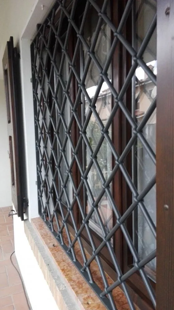 Inferriata fissa in ferro con maglia a rombi davanti a una finestra con scuroni in legno.