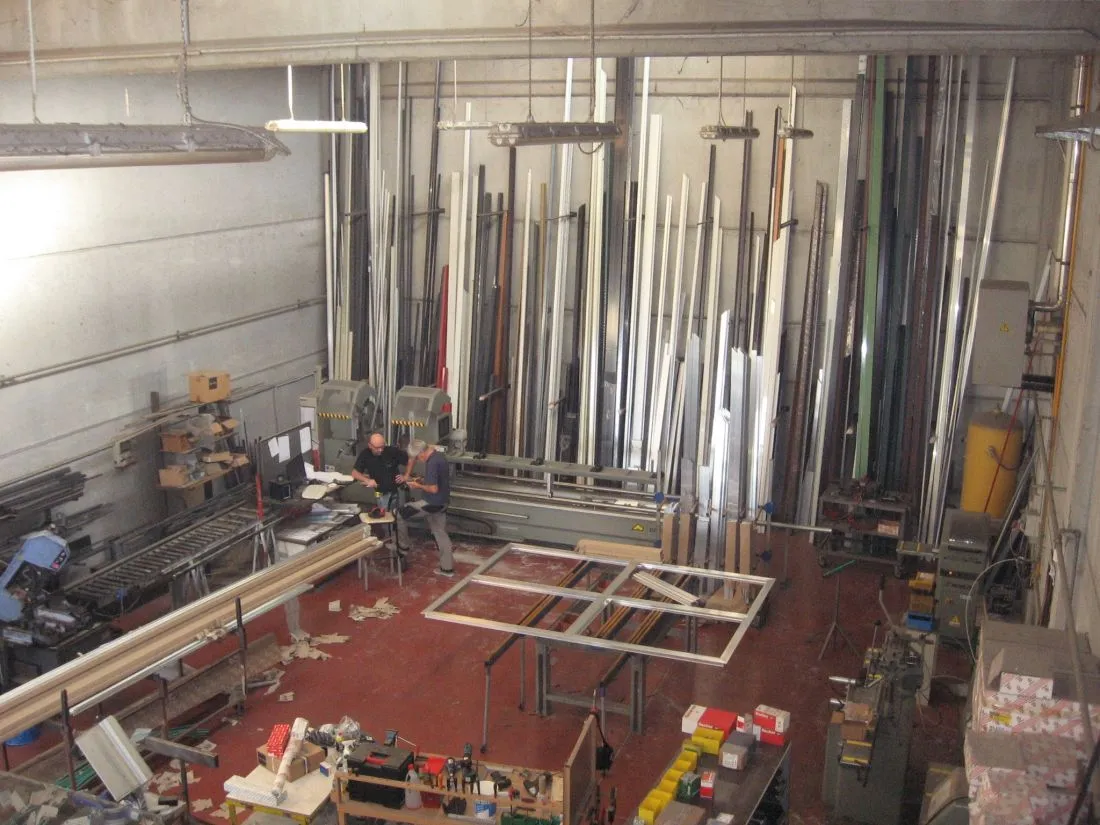 Interno dell'officina con barre di profili in alluminio in verticale e un telaio in lavorazione sul banco.