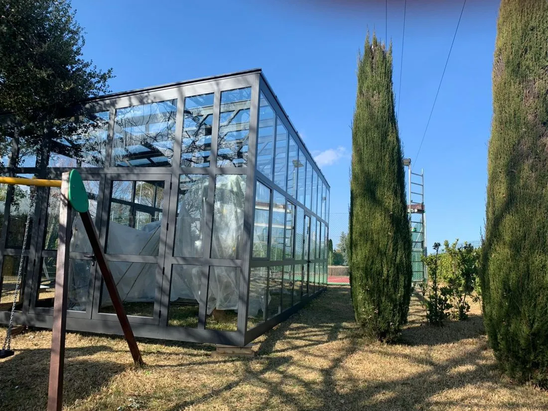 Serra in alluminio e vetro in un giardino con cipressi, con piante coperte al riparo.